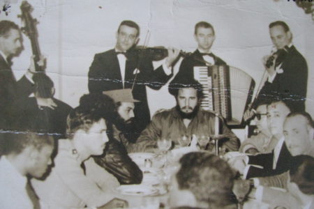 Fidel Castro dining out with his government entourage and military guards, with a small band of accordion and string players serenading him, 1961