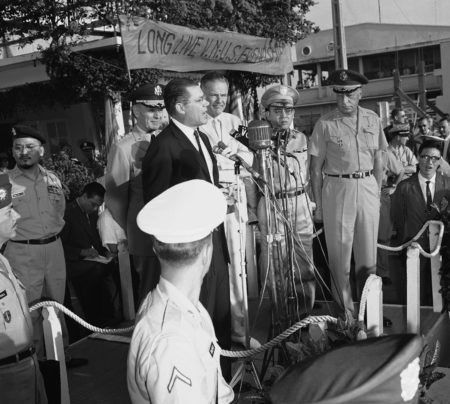 U.S. Defense Secretary Robert McNamara addresses welcome crowd at Saigon airport on March 8, 1964 under "long live Vietnamese US friendship" banner. Left chairman Joint Chiefs of Staff general Marwell D. Taylor, right Ambassador Henry Lodge and Vietnam Defense Minister General Tran Thien Khiem.
