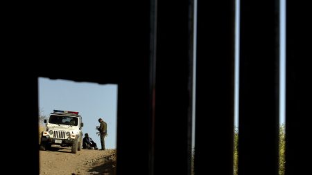 A U.S. Border Patrol agent questions a man in Nogales, Ariz., seen through a hole in a metal fence marking the border between the U.S. and Mexico, in 2007.