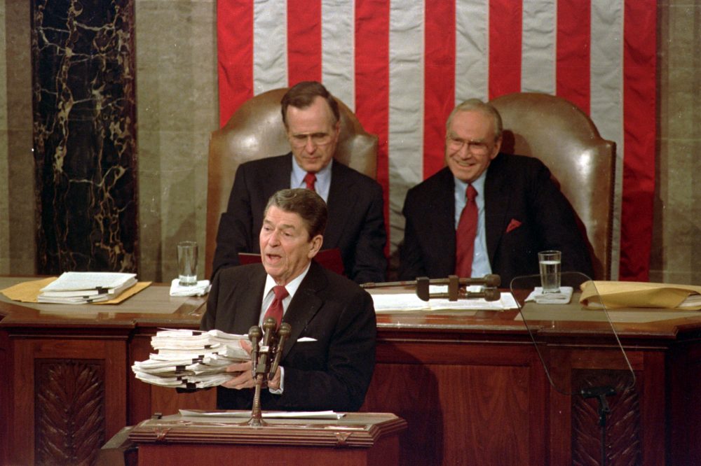 During his 1988 State of the Union address, President Ronald Reagan holds up a 14-pound continuing resolution for the budget, part of a total package weighing 43-pounds, which the president said was two months late from Congress. Vice President George H.W. Bush (left) and House Speaker James Wright of Texas listen behind him.
