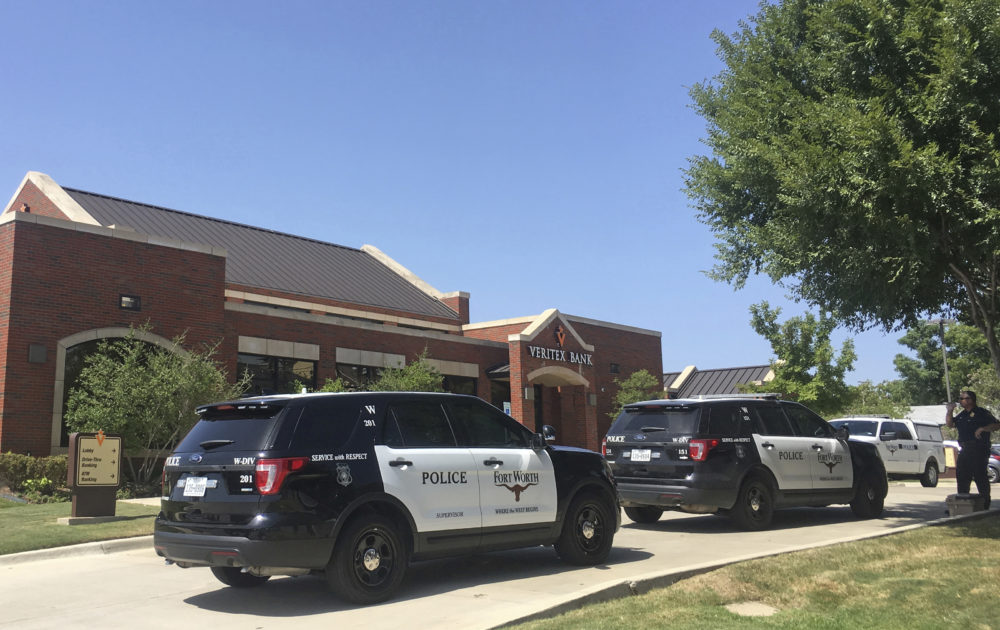 A Fort Worth police officer and vehicles are seen outside a Veritex Bank in Fort Worth, Texas, Thursday, July 19, 2018, following a shooting which took place inside the bank. Police are searching for two armed suspects after three bank employees were shot but are expected to survive. Officers initially responded to a robbery call Thursday, but Sgt. Chris Britt says investigators are trying to determine whether the shootings were part of a robbery attempt "or what exactly was going on." 