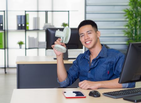 Asian young man's sitting at an office chair holding a small plastic fan blowing to his face because hot weather. He's looking at camera and smile with relaxation after use a fan in studio workplace