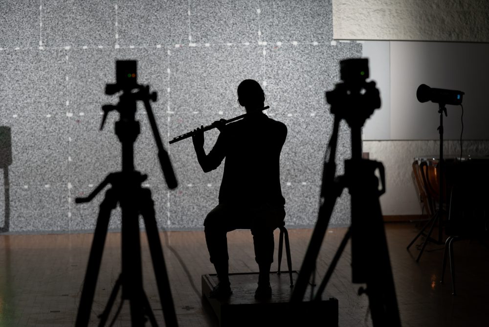 Musicians perform in front of a highly-textured background. Microdroplets from their playing will make the background appear to move in the video captured by the team's camera. 