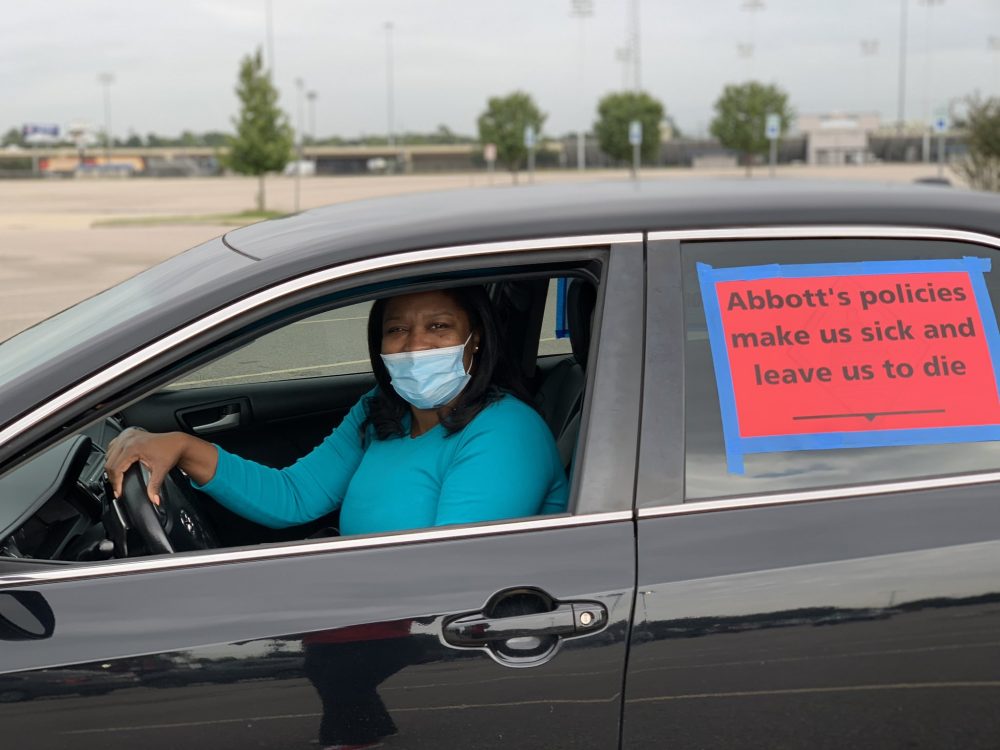 An educator wears a mask and sits in her car with a sign on the window that says "Abbott's policies make us sick and leave us to die."