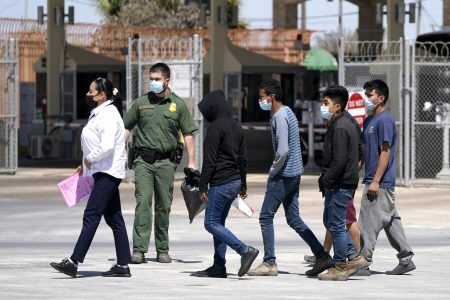 Migrants, who were caught trying to sneak into the United States, are led by a U.S. Customs and Border Protection agent, second from left, at the McAllen-Hidalgo International Bridge while being deported to Reynosa, Mexico, Thursday, March 18, 2021, in Hidalgo, Texas. A surge of migrants on the Southwest border has the Biden administration on the defensive. The head of Homeland Security acknowledged the severity of the problem Tuesday but insisted it's under control and said he won't revive a Trump-era practice of immediately expelling teens and children. (AP Photo/Julio Cortez)