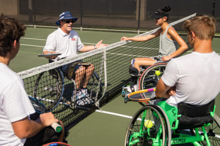 youth tennis players in sporty wheelchairs gather around a coach giving instruction on a green tennis court