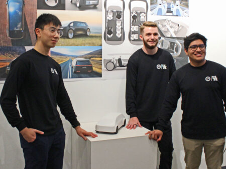 Three students wearing black shirts pose with a small white 3D printed model of an electric vehicle. Printed computer renderings of the design are behind them on a wall.