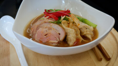 A neatly plated bowl of dumplings, pork, brother and noodles sits on a wooden tray with chopsticks and a spoon.