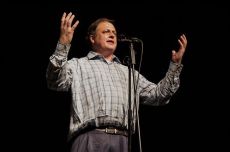 Houston storyteller Greg Audel standing on a dark stage with his arms raised.
