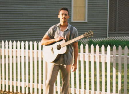 Musician Micah Edwards poses with a guitar in front of a house.