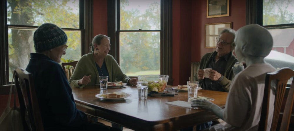 Photo of four people sitting around a table--one is an alien from space