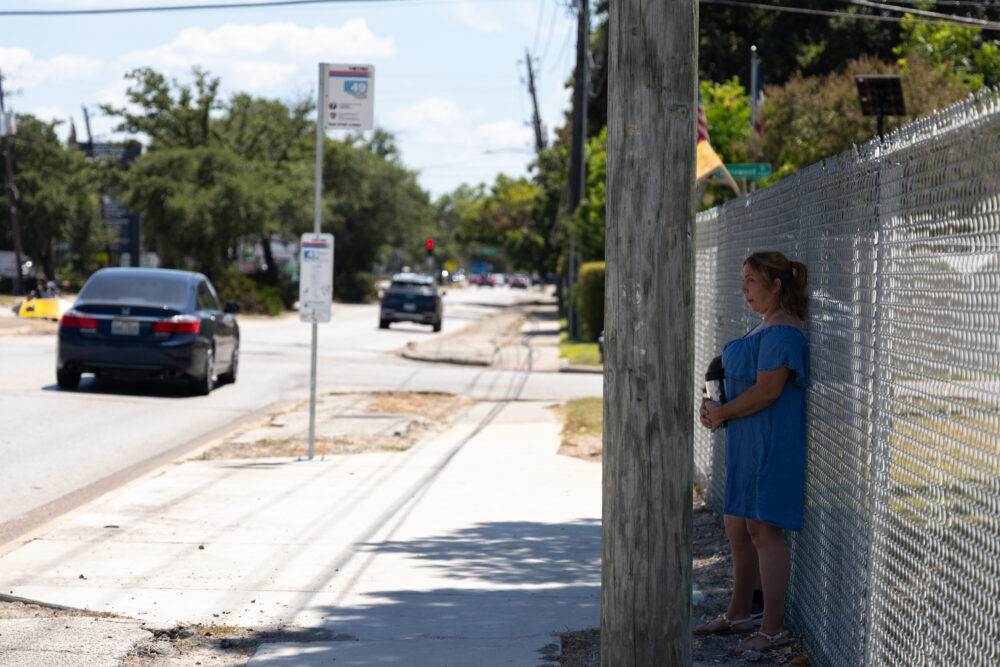 A woman waits in the shade away from a bus stop until the bus arrives.