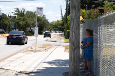 A woman waits in the shade away from a bus stop as she waits for her bus to arrive.