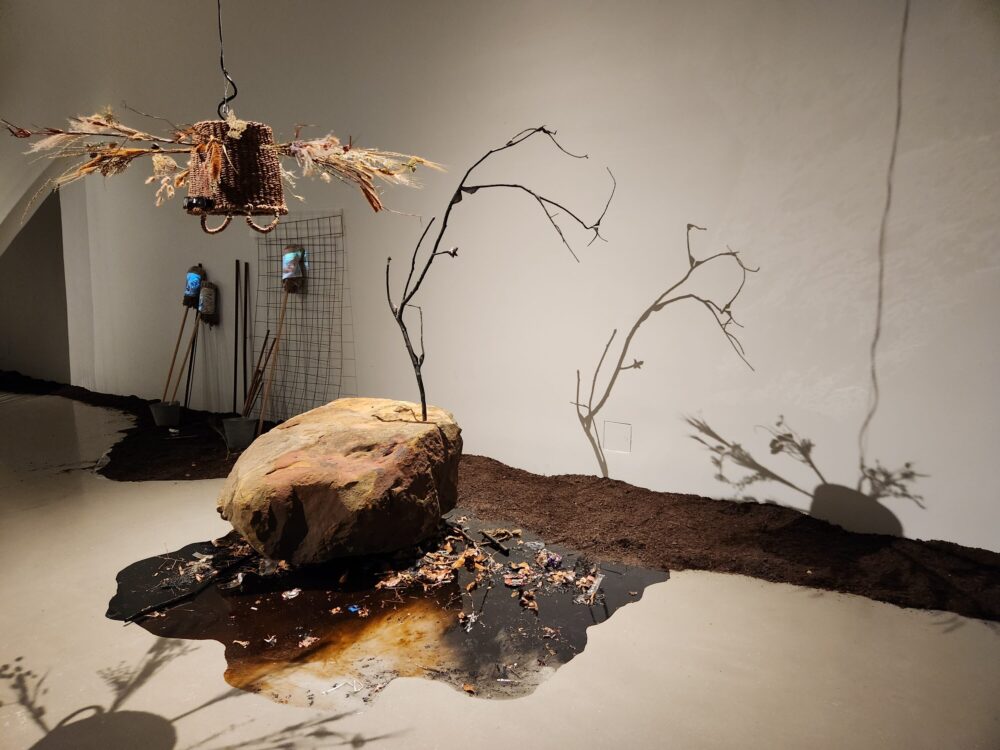 Photo of art exhibit with a fake oil spill, a rock, and a tree