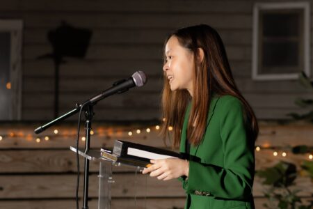 Elizabeth Hsu, in a green suit, stands in front of a microphone to speak at her inauguration as Houston Youth Poet Laureate in November 2023.