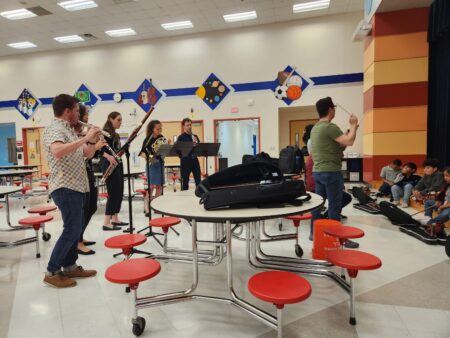 Professional musicians perform with elementary school students in a school cafeteria.
