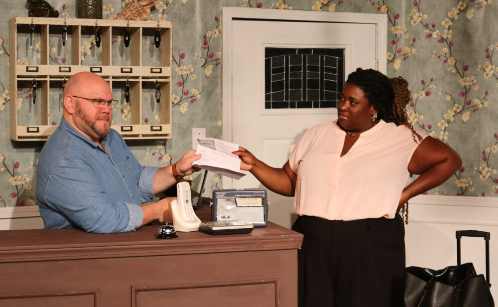 A man behind a desk talks to a woman who is holding a newspaper