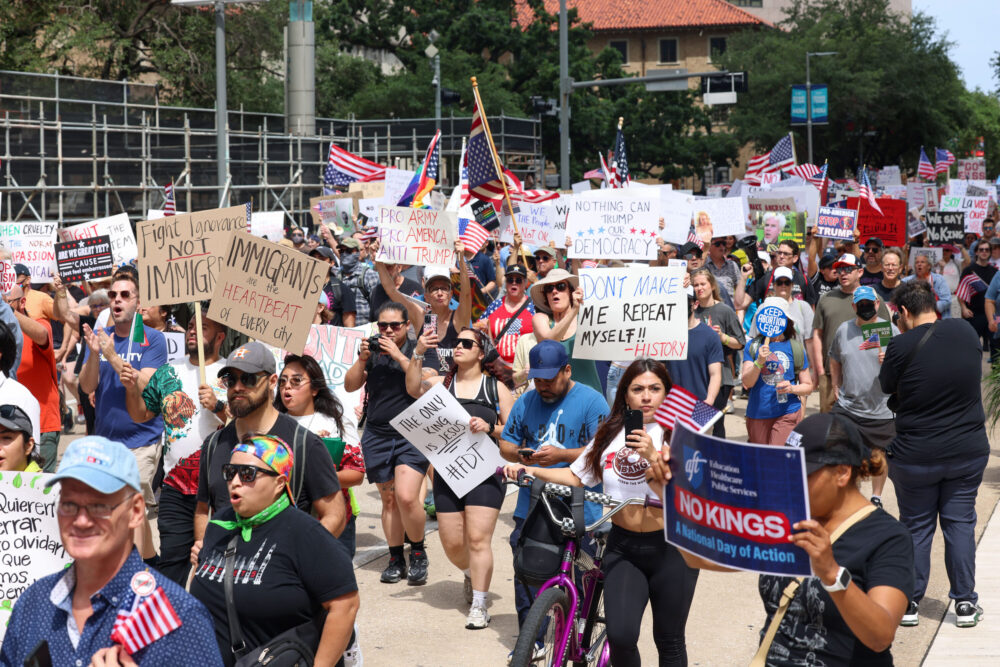 Berjalanlah para pengunjuk rasa sambil memelihara poster di pusat kota Houston pada hari Sabtu, 14 Juni 2025. (Kredit foto: Rob Salinas/Houston Public Media)